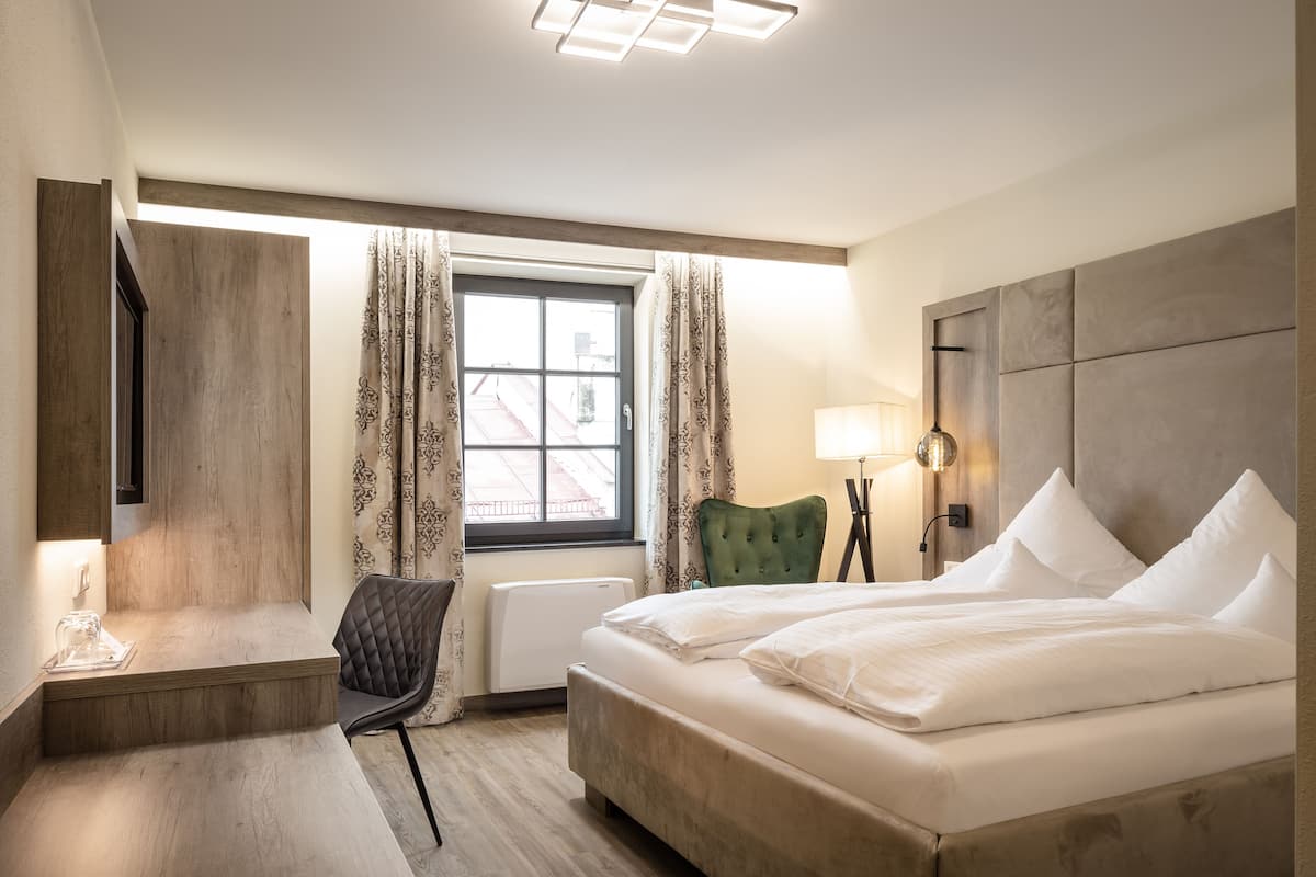 Une chambre d’hôtel avec un lit, un bureau, une chaise, une fenêtre avec des rideaux et une lampe.