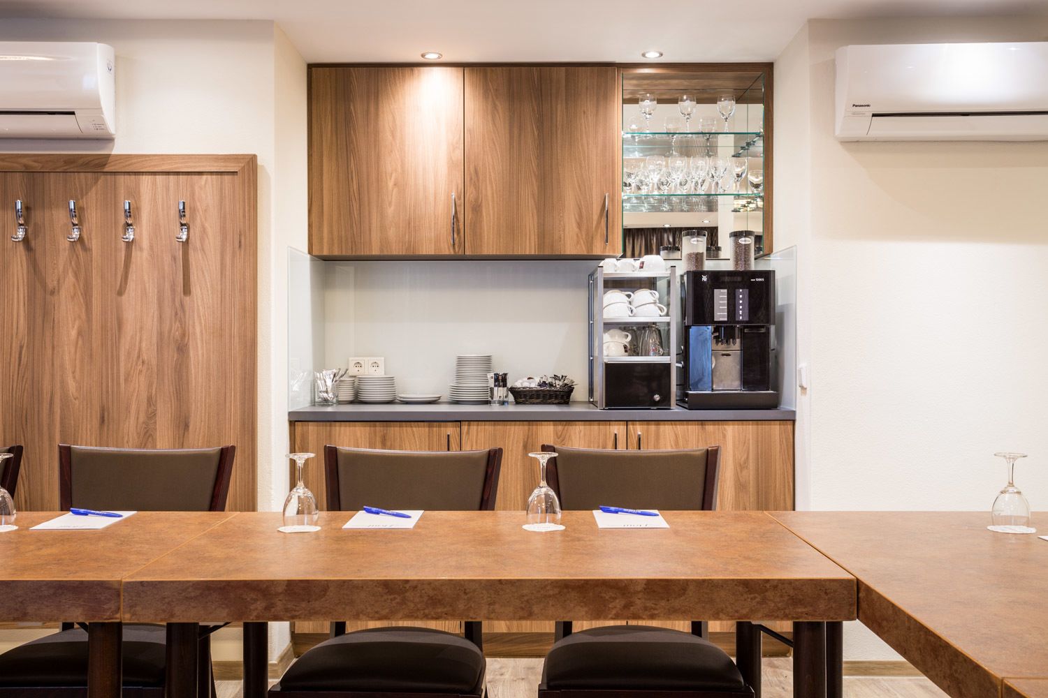 A modern kitchen with wooden cabinets, a black coffee machine, and a glass display case with wine glasses.
