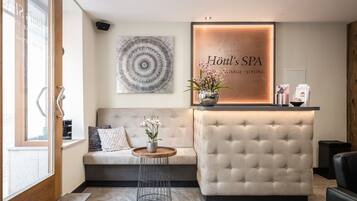 A reception area with a tufted sofa, a round wooden table with a vase of flowers, a wall-mounted sign for Höttl's SPA, and a large abstract artwork.