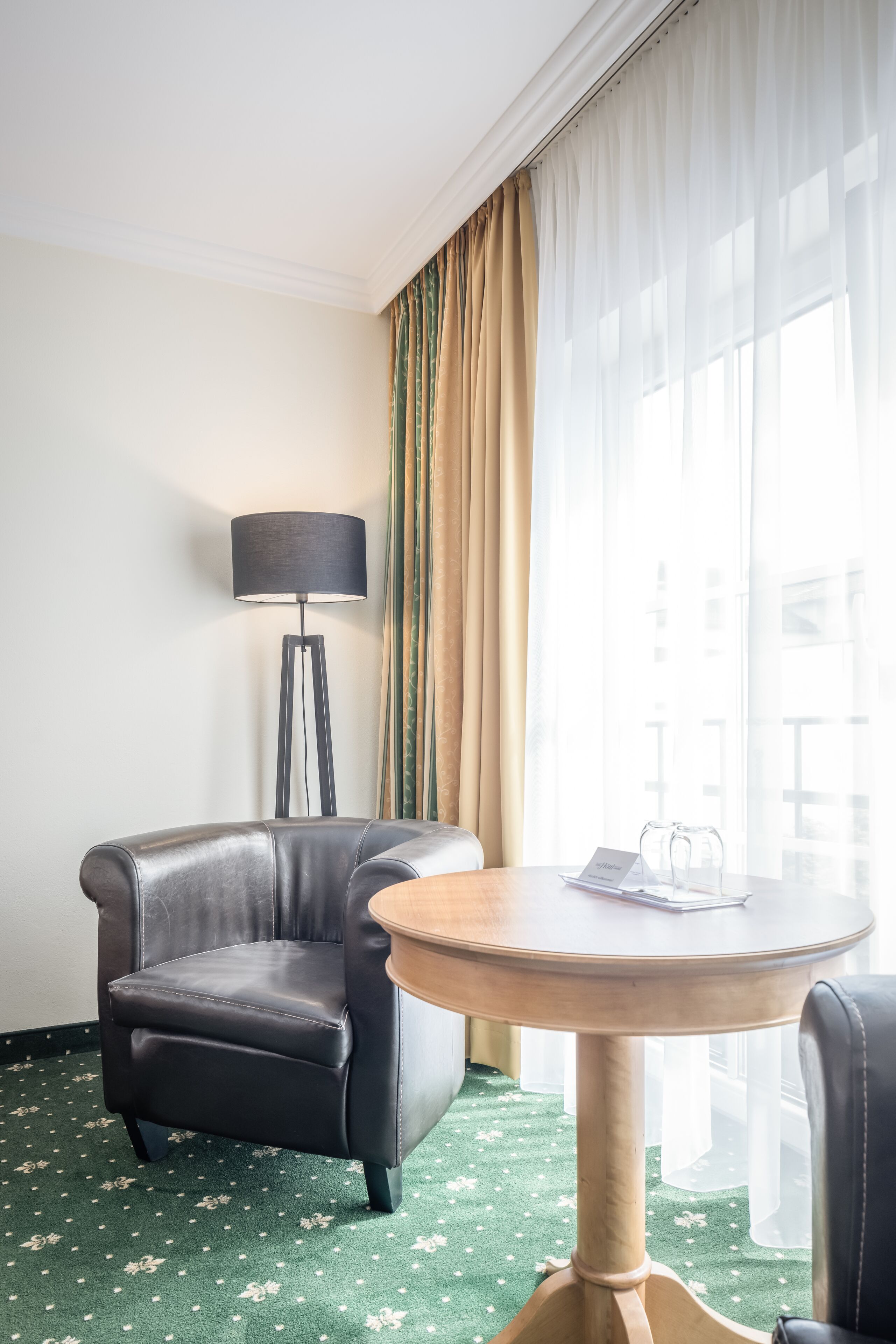 A hotel room with a leather armchair, a round wooden table, a floor lamp, sheer curtains, and a view of the city through large windows.