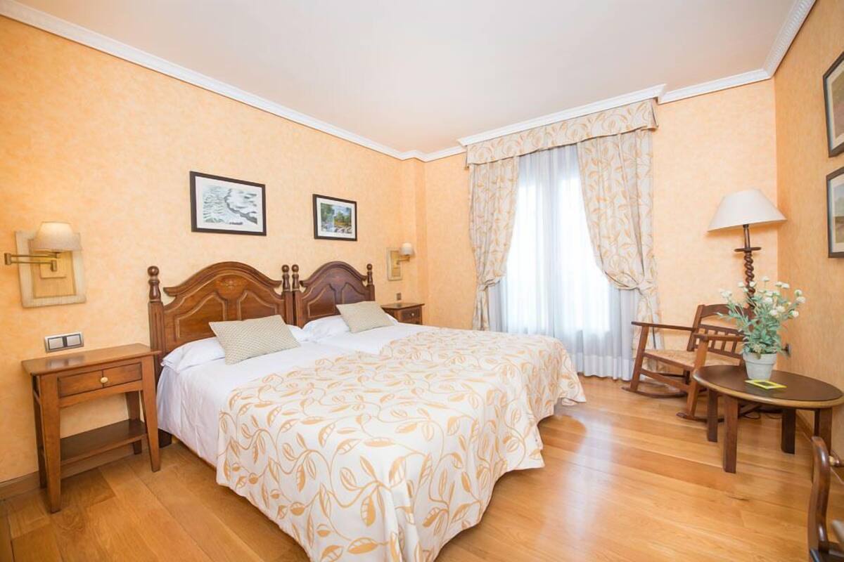 Ett hotellrum med en säng i trä, två sängbord, en stol, ett litet bord med en lampa och ett fönster med gardiner.
