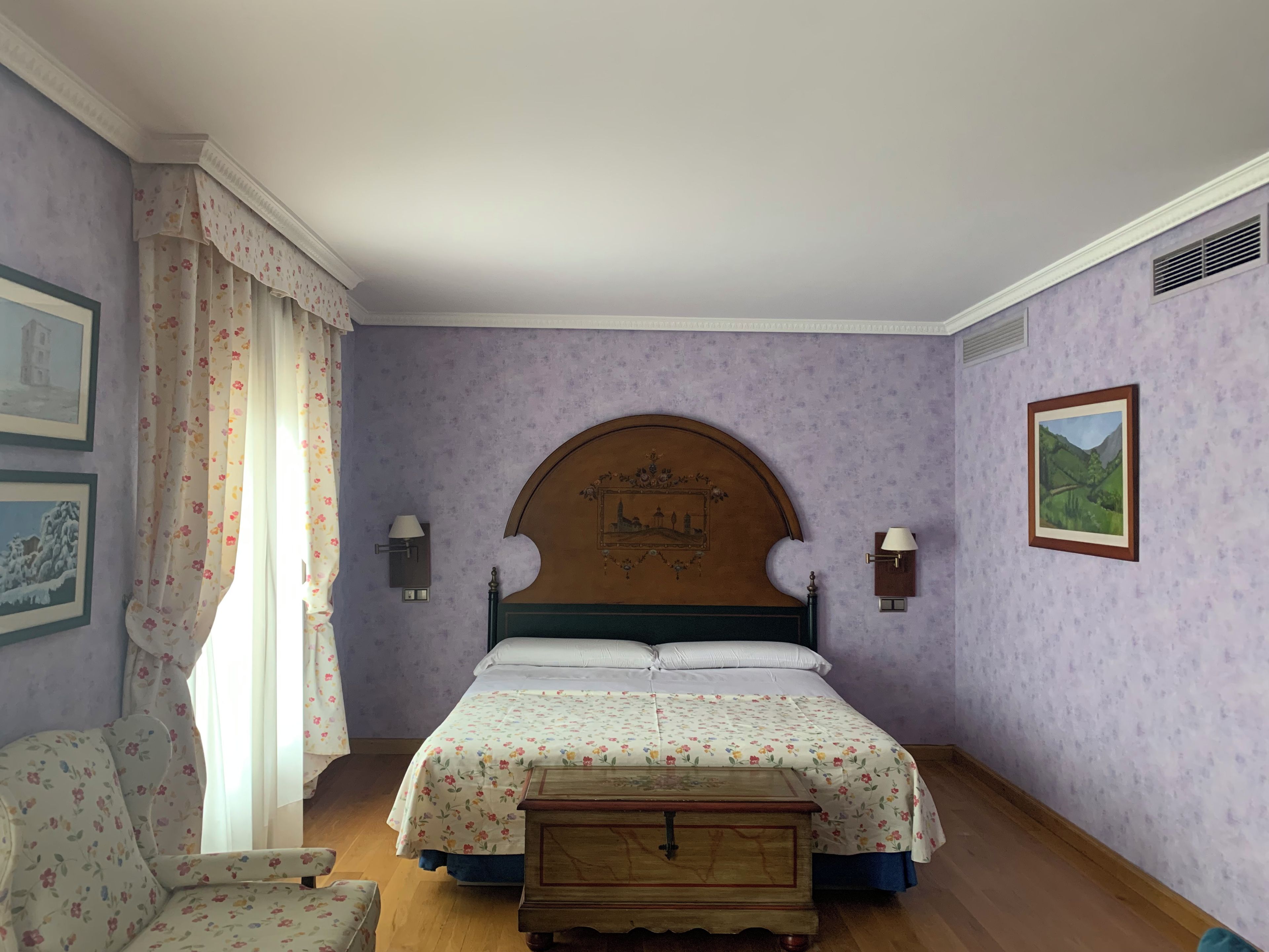 Un dormitorio con cama, mesita de noche, una silla y dos lámparas de pared.
