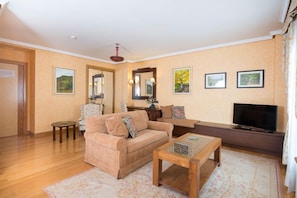 Ein Wohnzimmer mit Sofa, Couchtisch und Fernseher. An den Wänden hängen Bilder in Rahmen, und über dem Sofa befindet sich ein Spiegel.