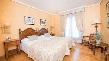 Una habitació d'hotel amb un llit de fusta, dues taules de nit, una cadira, una taula petita amb una làmpada i una finestra amb cortines.