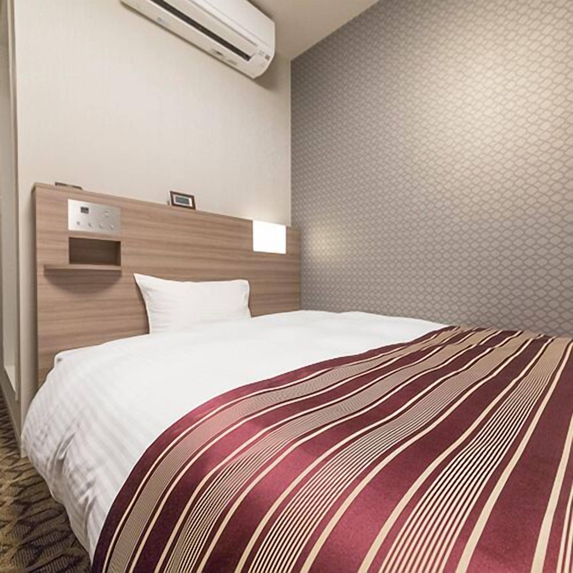 Ett hotellrum med en säng, en väggmonterad luftkonditioneringsanläggning, en liten hylla och en vägg med ett mönster.