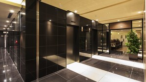 Un hall d’hôtel moderne avec un sol carrelé de noir, un écran numérique indiquant l’étage de l’ascenseur et un espace restaurant visible à travers des portes vitrées.