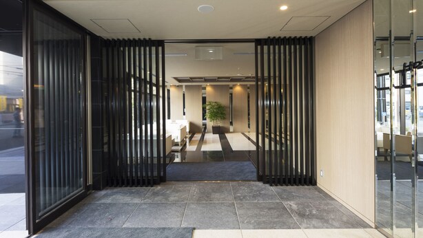 Une entrée d’hôtel moderne avec une porte en verre, une plante en pot et un espace salon visible à travers le verre.