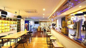Een restaurant met houten tafels en stoelen, een bar met flessen en een kassa, en een muur met schappen vol toevoegingen en een Pepsi-bord.