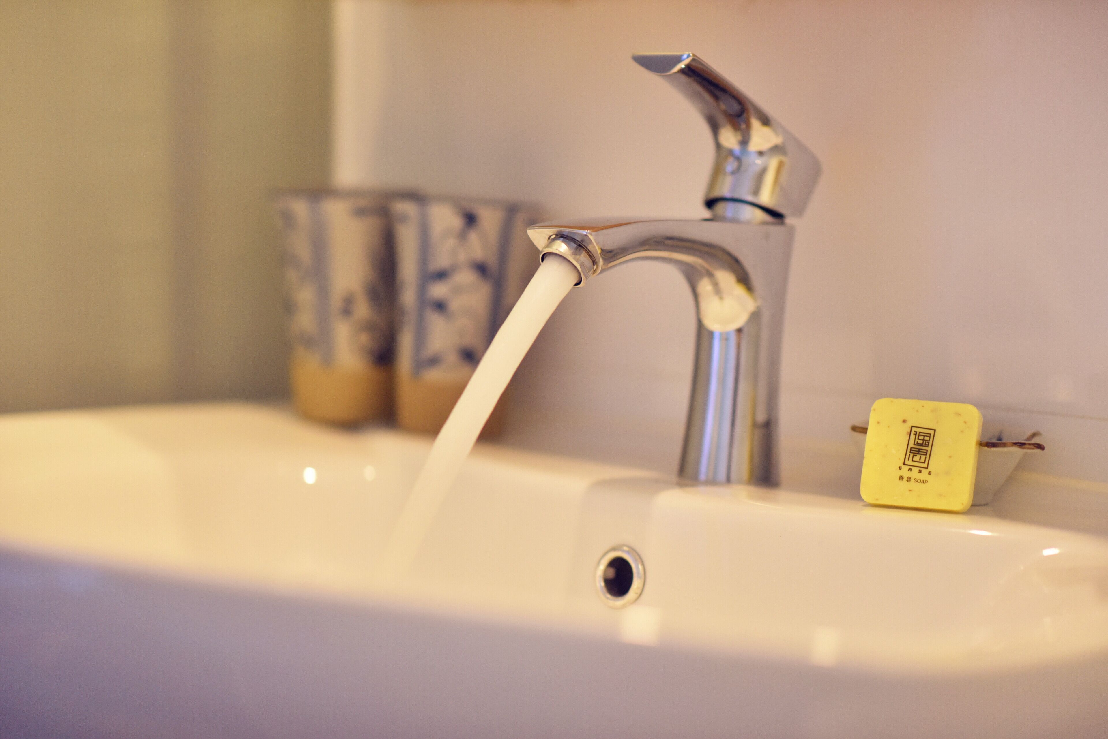 A bathroom sink with a running faucet, a soap dispenser, and a decorative cup in the background.