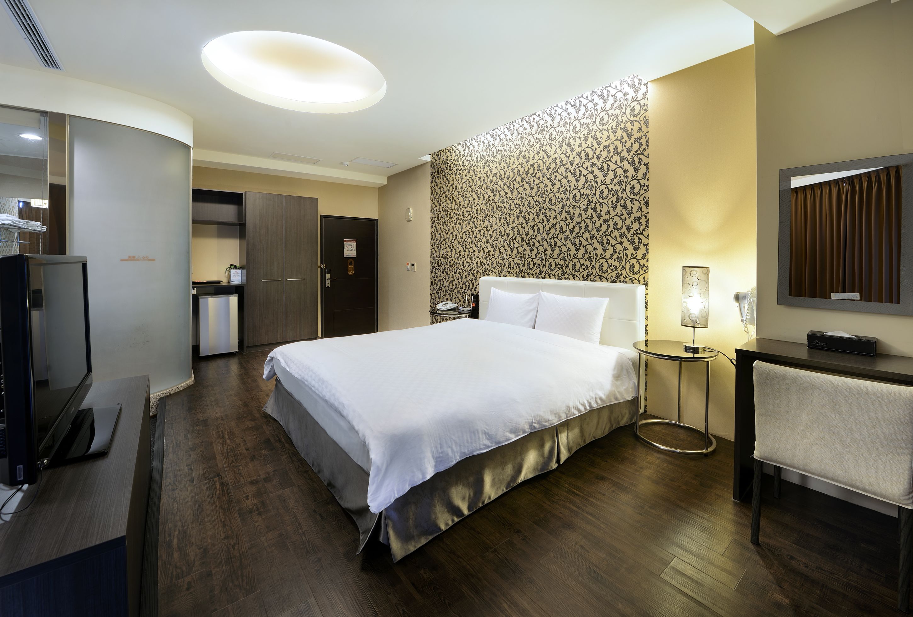 A hotel room with a large bed, a desk, a chair, a TV, a small table with a lamp, and a patterned wall.