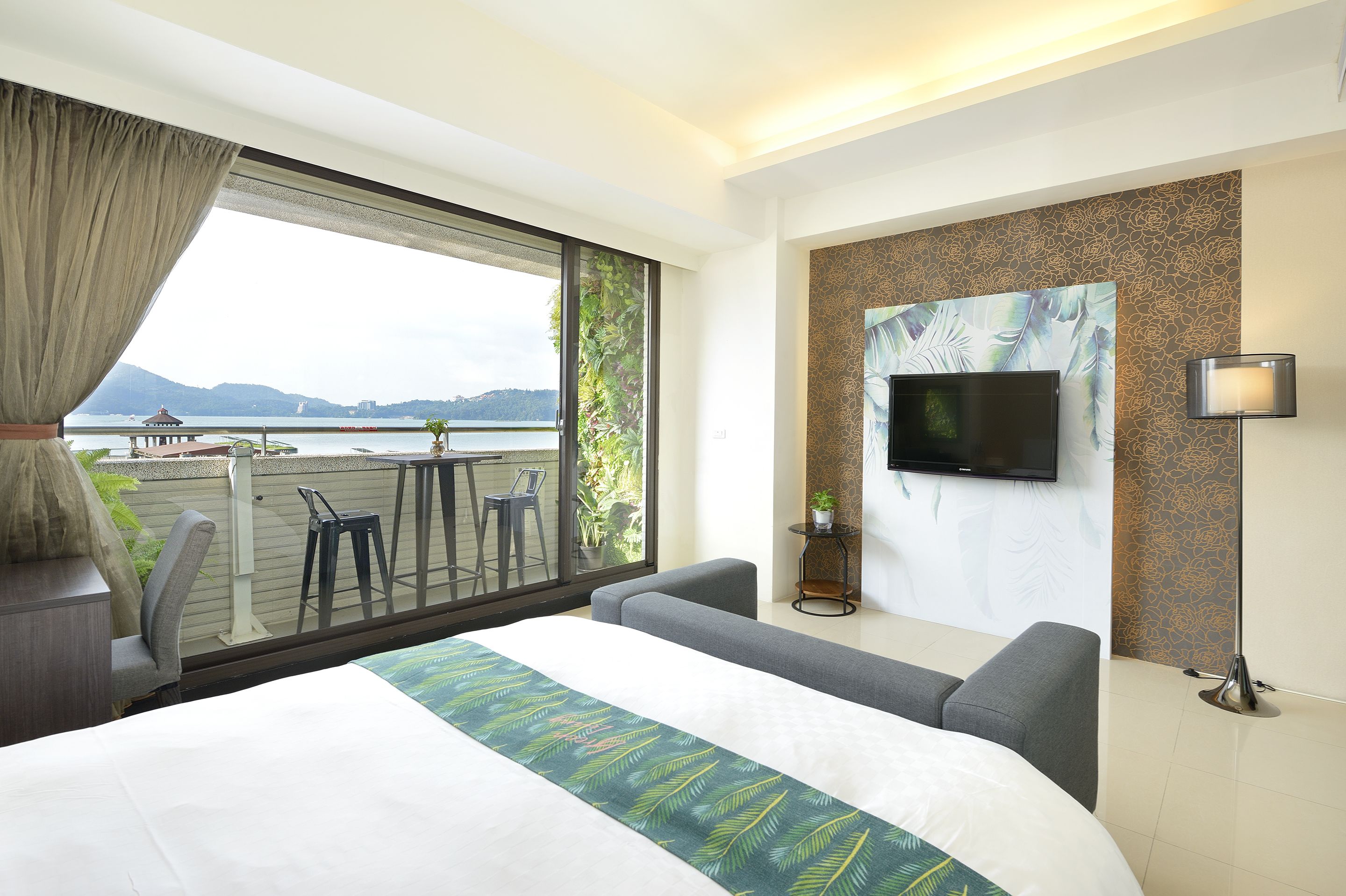 A hotel room with a large bed, a TV mounted on the wall, a balcony with a view of the water and mountains, and a seating area with a table and chairs.