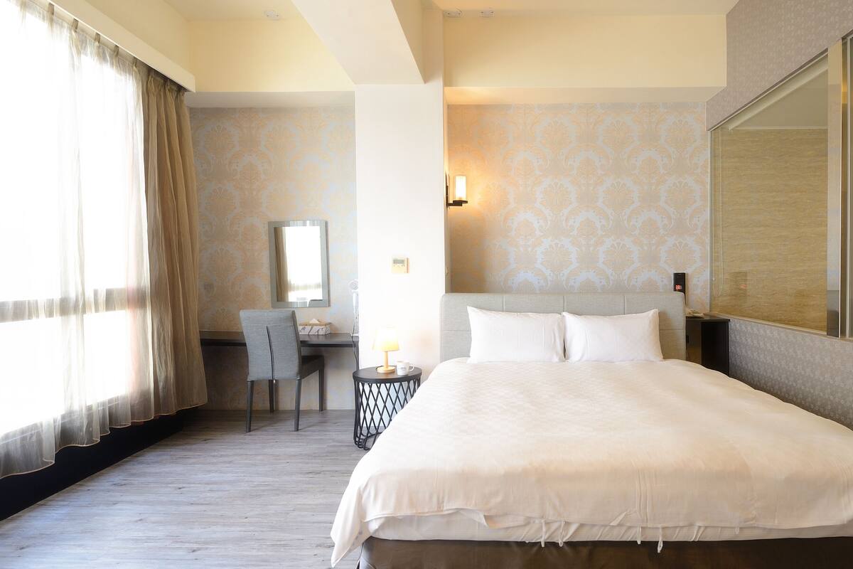 Habitación de hotel con una cama grande, un escritorio con una silla, una mesita con una lámpara y un espejo grande en la pared.