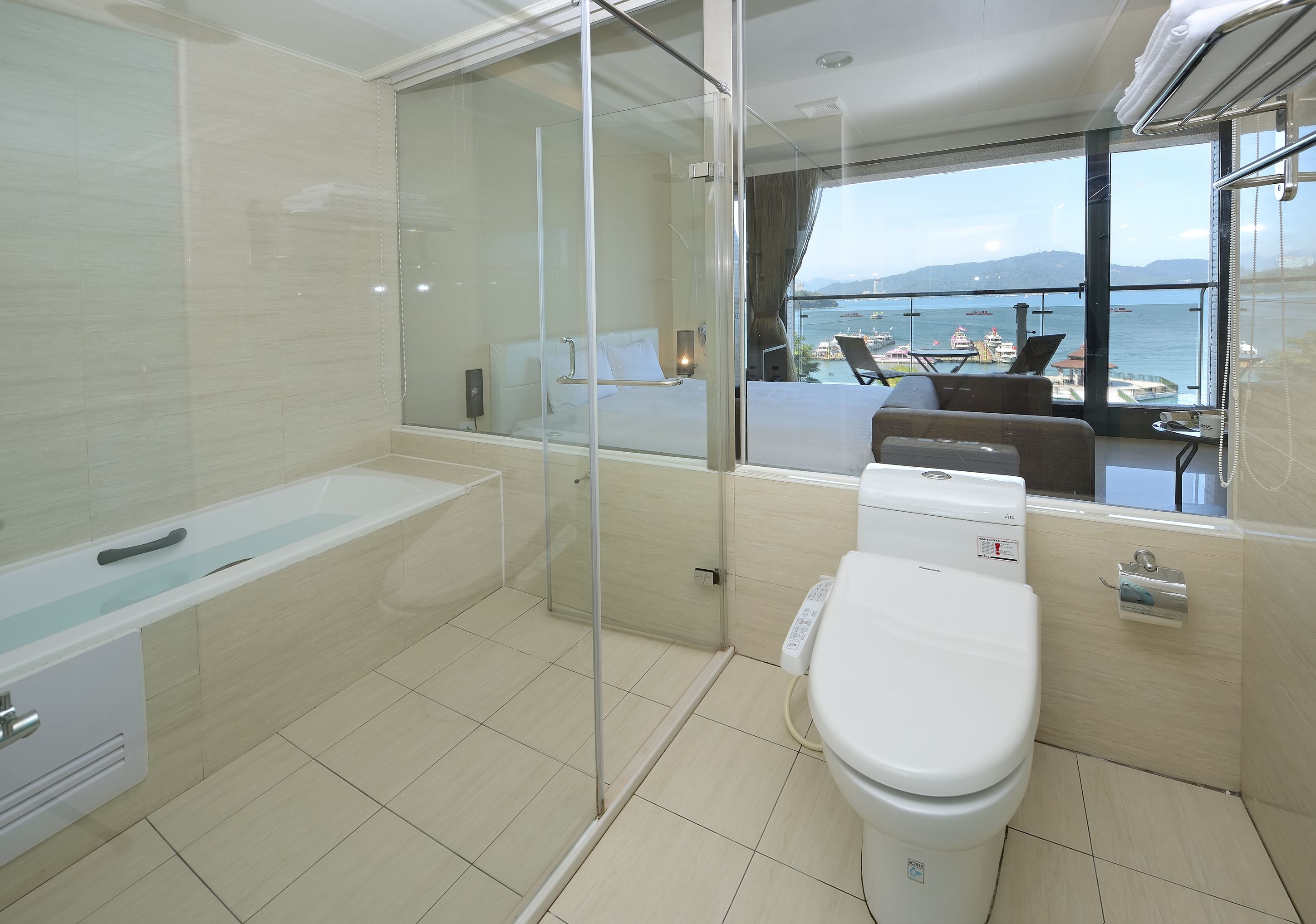 A modern bathroom with a bathtub, toilet, and glass shower enclosure. A large window offers a view of the sea and boats.