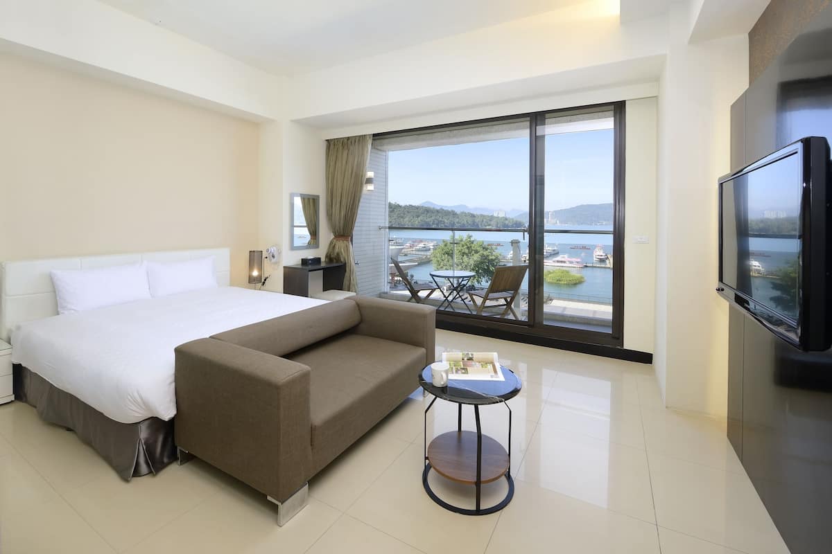 Una habitación de hotel moderna con una cama grande, un sofá, una mesita, un televisor y un balcón con vistas al mar y a las embarcaciones.