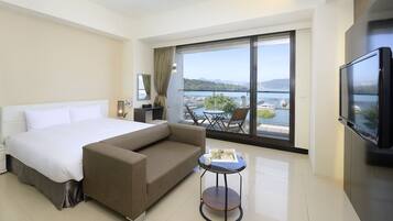 A modern hotel room with a large bed, a sofa, a small table, a TV, and a balcony with a view of the sea and boats.