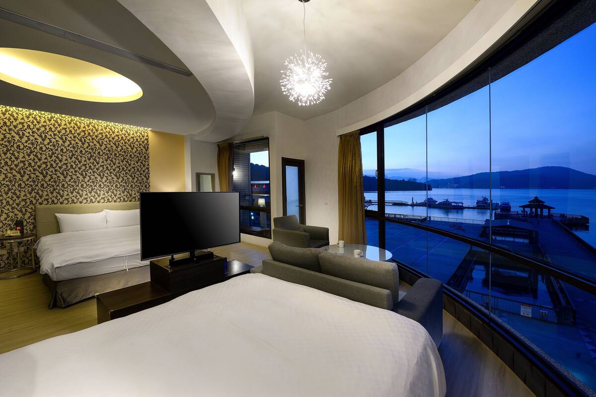 Una habitación de hotel moderna con una cama grande, un televisor de pantalla plana, un sofá y un balcón con vistas al agua y al muelle.