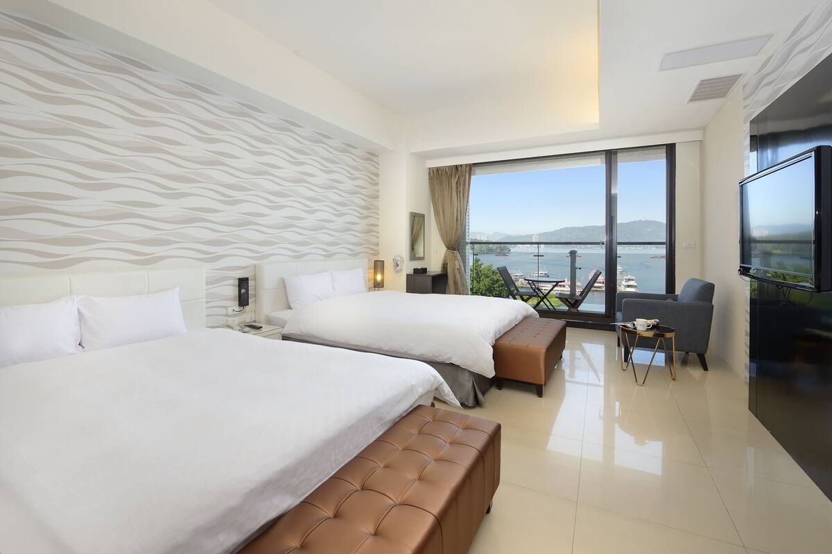 Una habitación de hotel moderna con dos camas, un ventanal con vistas al mar y un balcón con una mesa y sillas.