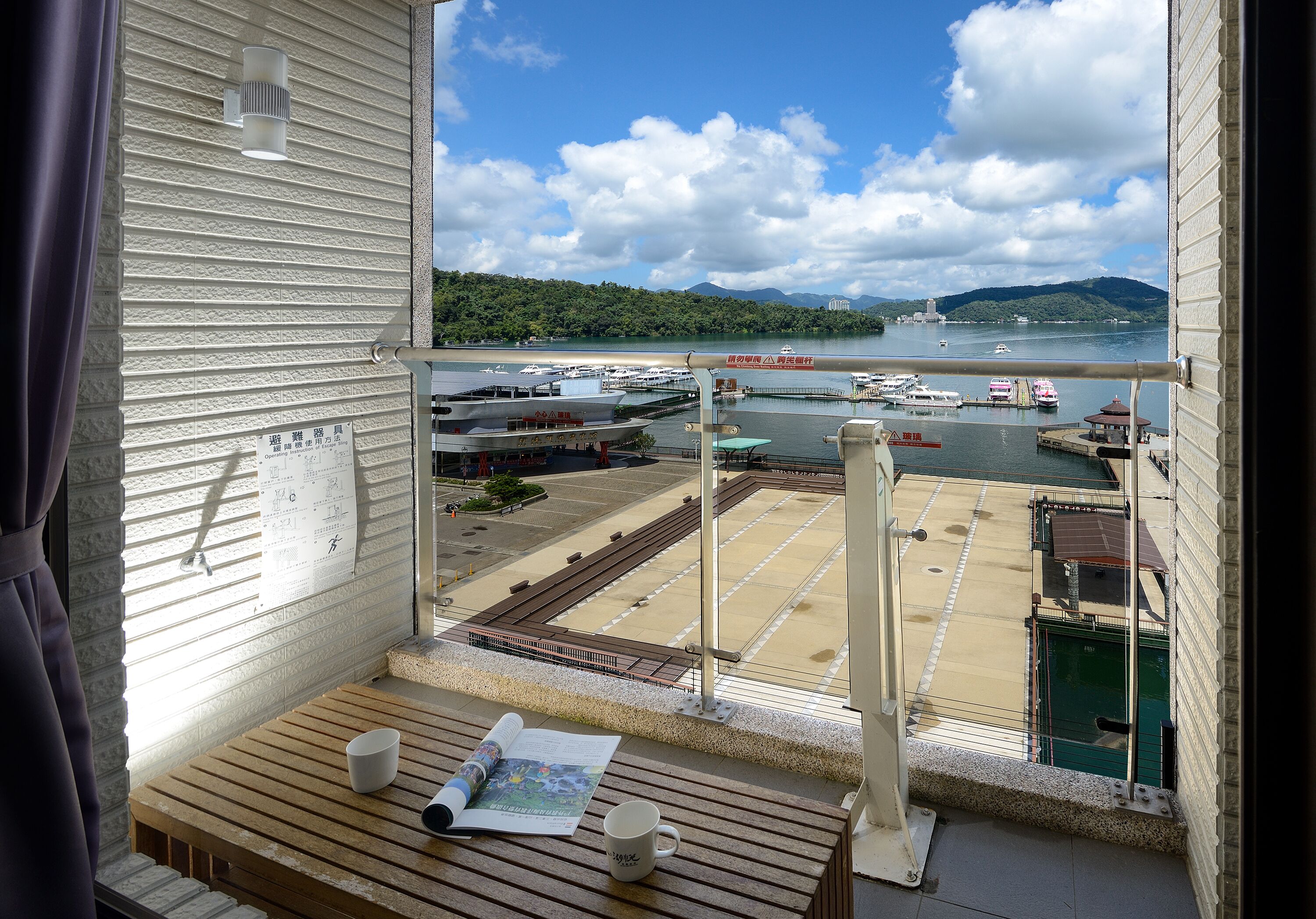 A balcony with a view of a marina, a wooden table with a cup and a magazine, and a window with blinds.