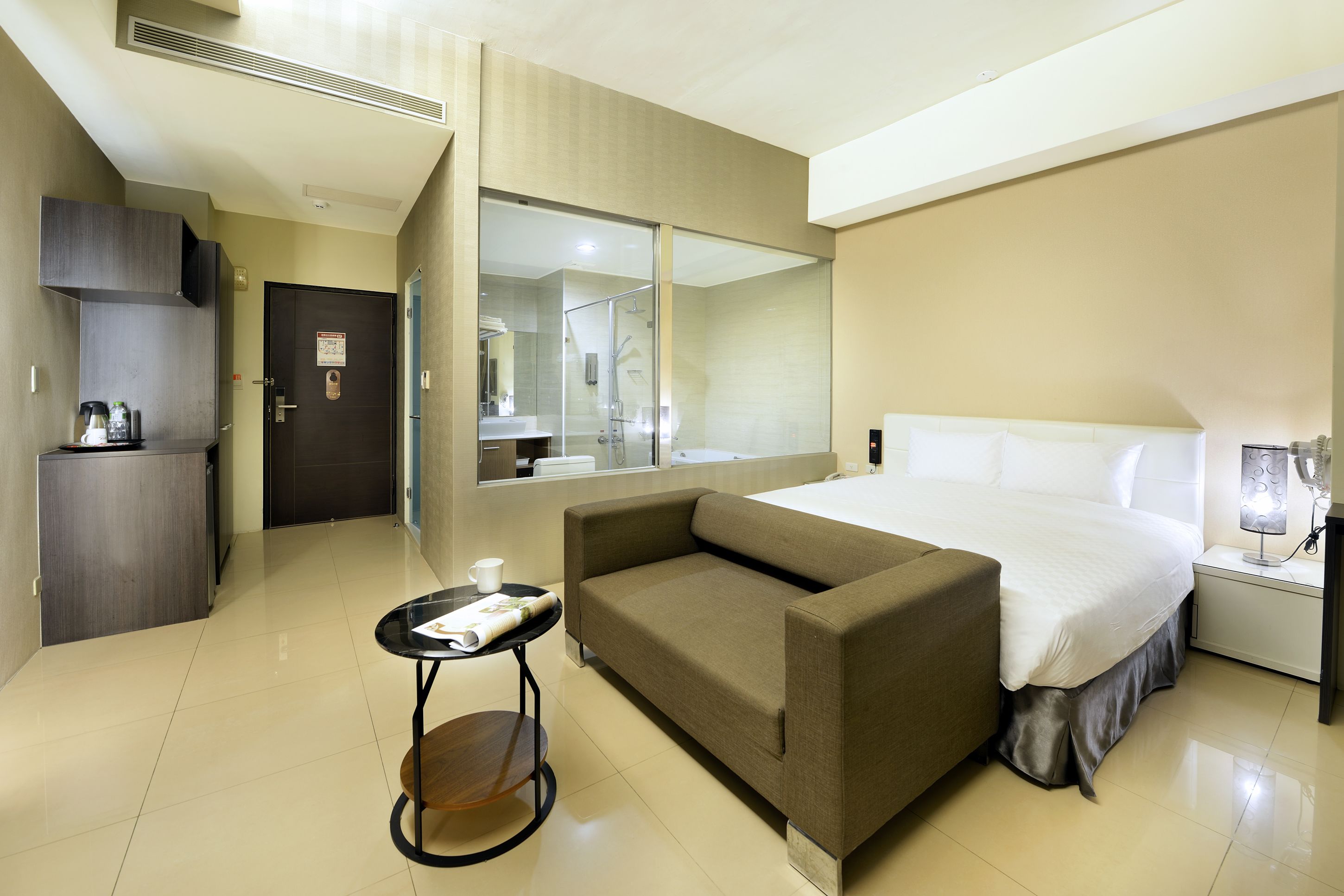 A hotel room with a bed, a sofa, a small table, a mirror, a wardrobe, and a bathroom visible through a glass door.