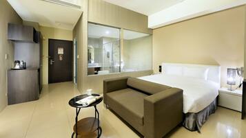 A hotel room with a bed, a sofa, a small table, a mirror, a wardrobe, and a bathroom visible through a glass door.