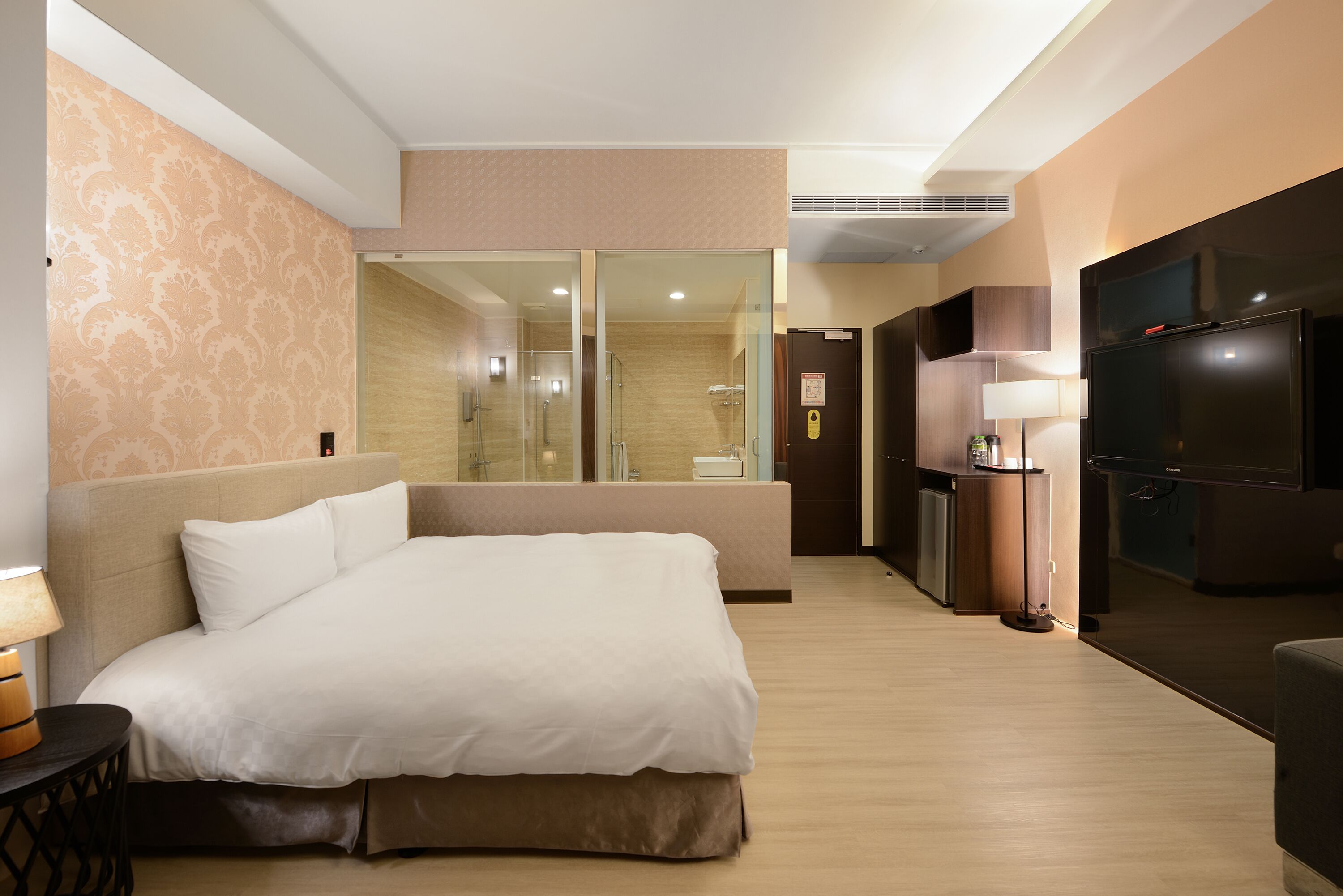 Habitación de hotel con una cama grande, televisión, escritorio y baño visible a través de un panel de vidrio.