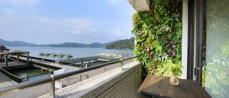 Un balcón con vistas a un lago y un muelle, una mesa de madera y un muro vertical cubierto de plantas.