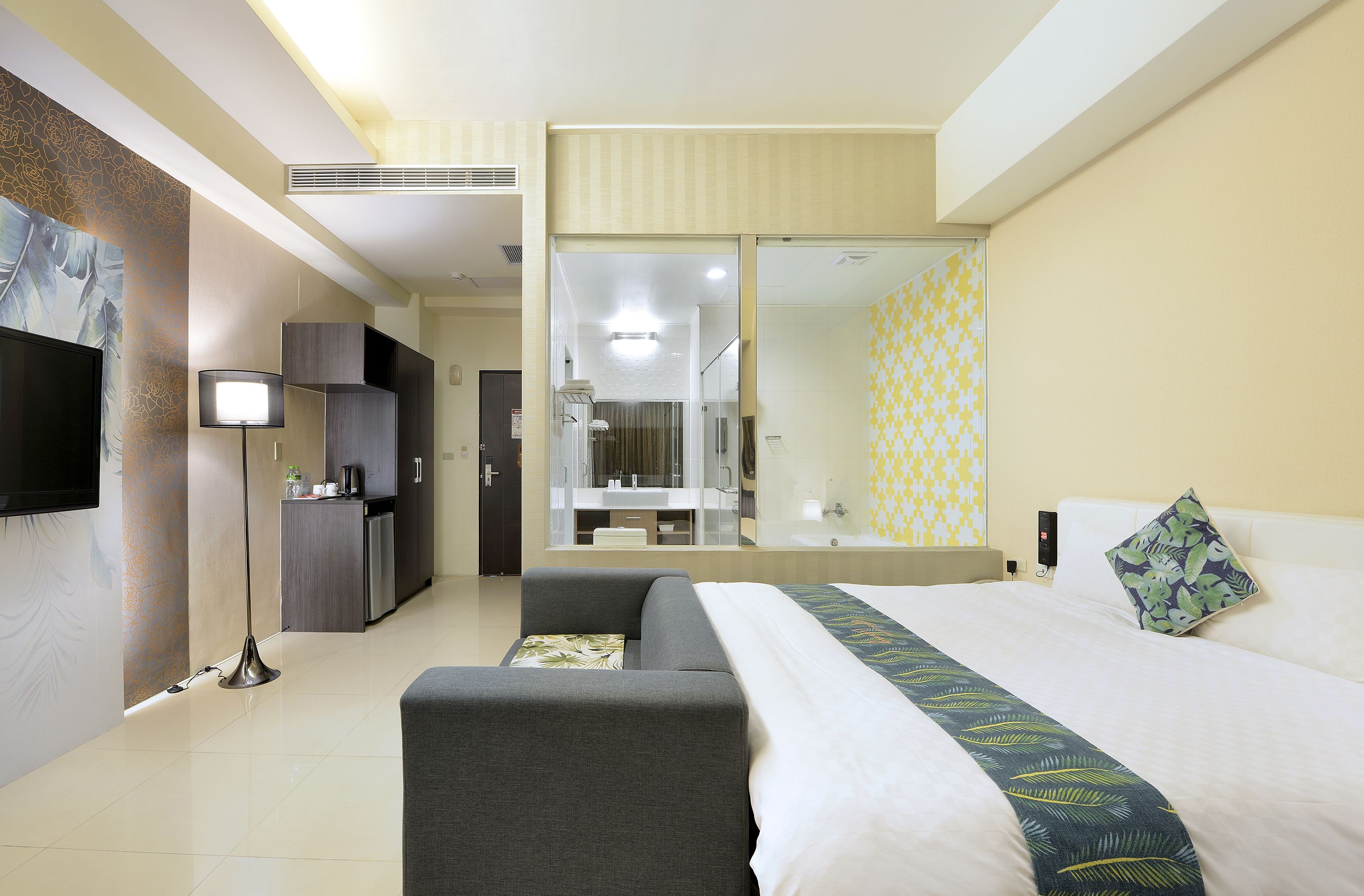 A hotel room with a bed, a sofa, a TV, a desk, and a bathroom visible through a glass door.