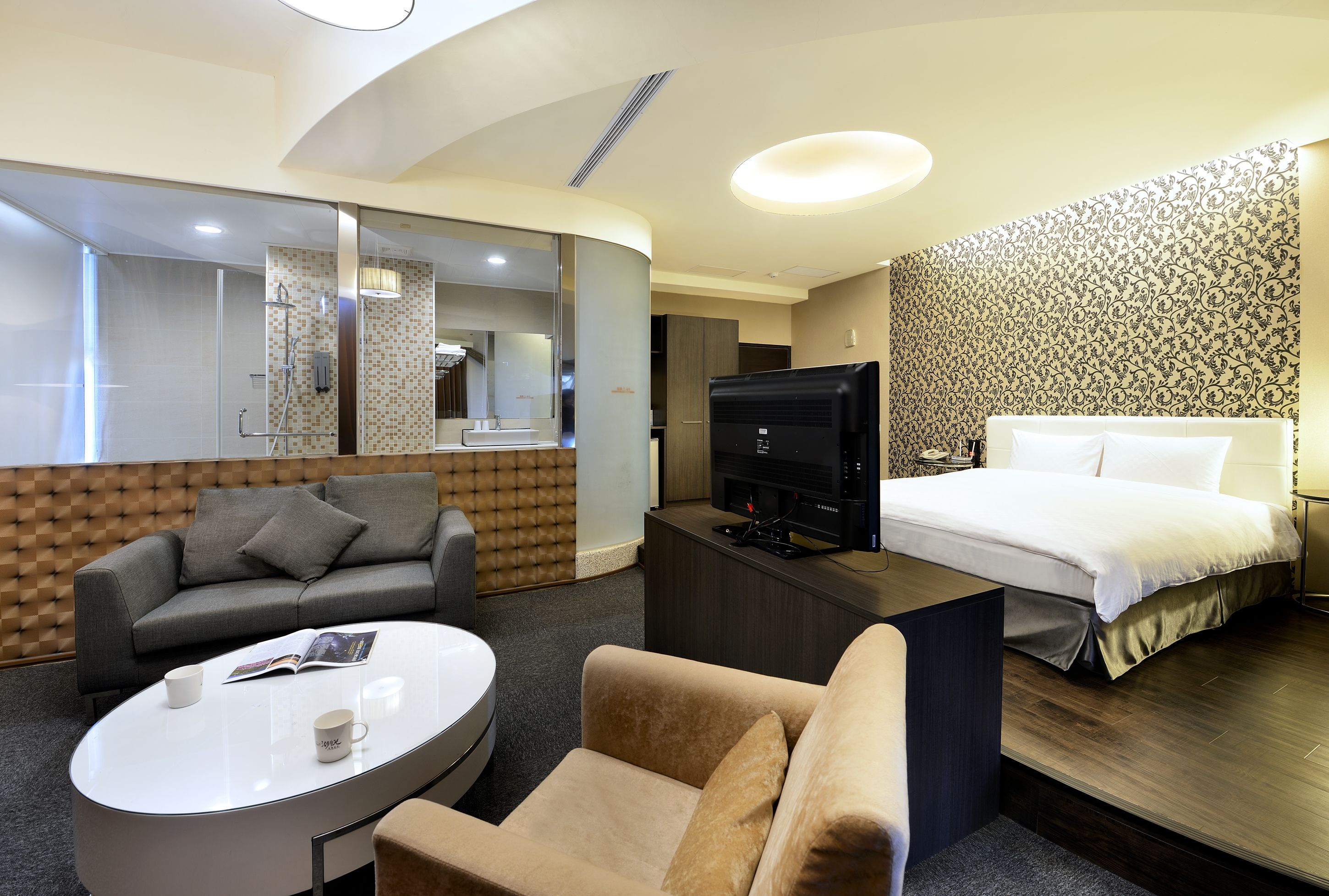 A modern hotel room with a sofa, a round coffee table, a TV, and a bed with a white duvet.