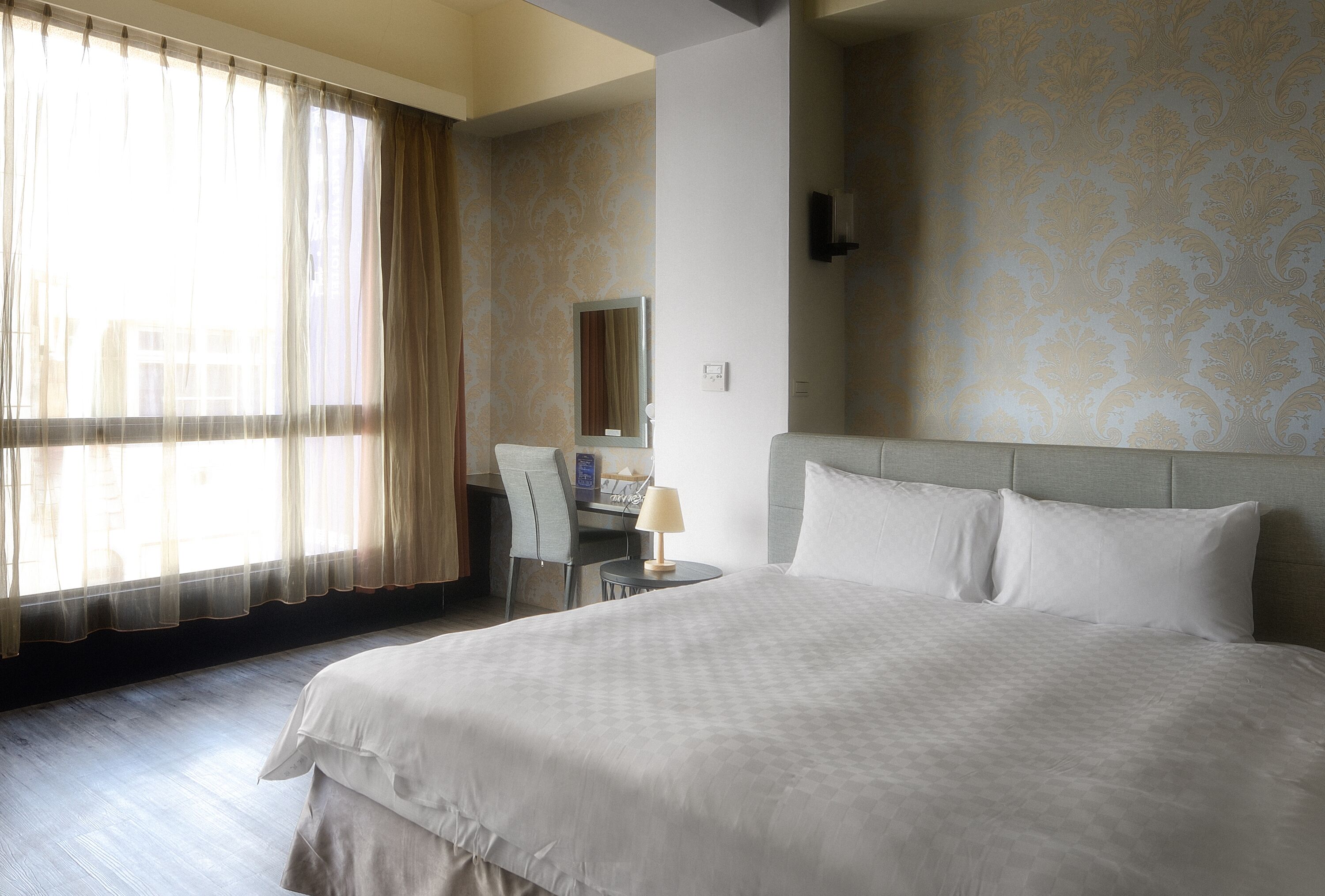 A hotel room with a large bed, a desk with a chair, a lamp, and a window with sheer curtains.