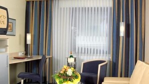 A hotel room with a bed, a desk, a chair, a small table with a vase of flowers, and a window with curtains. - Lowen-seckenheim (Mannheim)