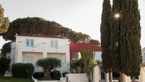 A two-story building with a white facade, a red awning, and a well-maintained garden with a large tree and shrubs. - Coluccia Hotel & Beach Club (Santa Teresa di Gallura)
