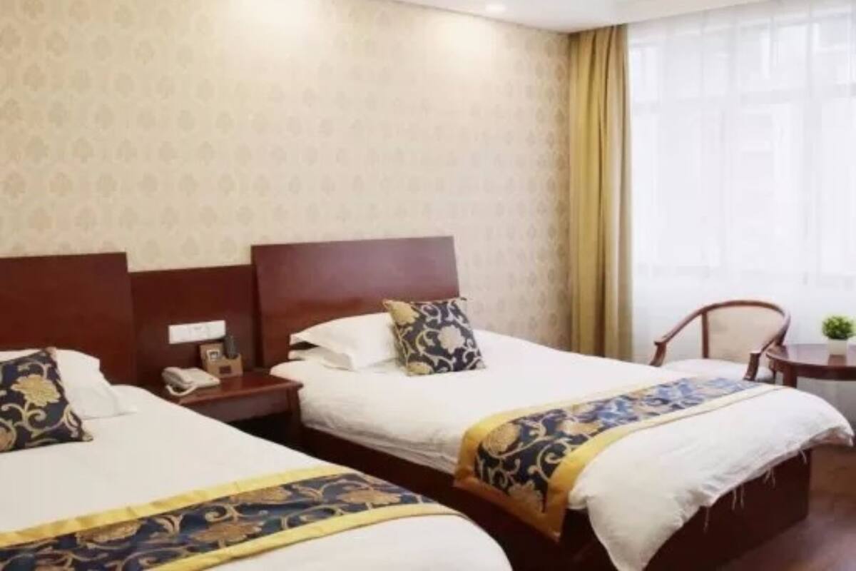 A hotel room with two beds, a wooden headboard, a small table with a chair, and a window with curtains.