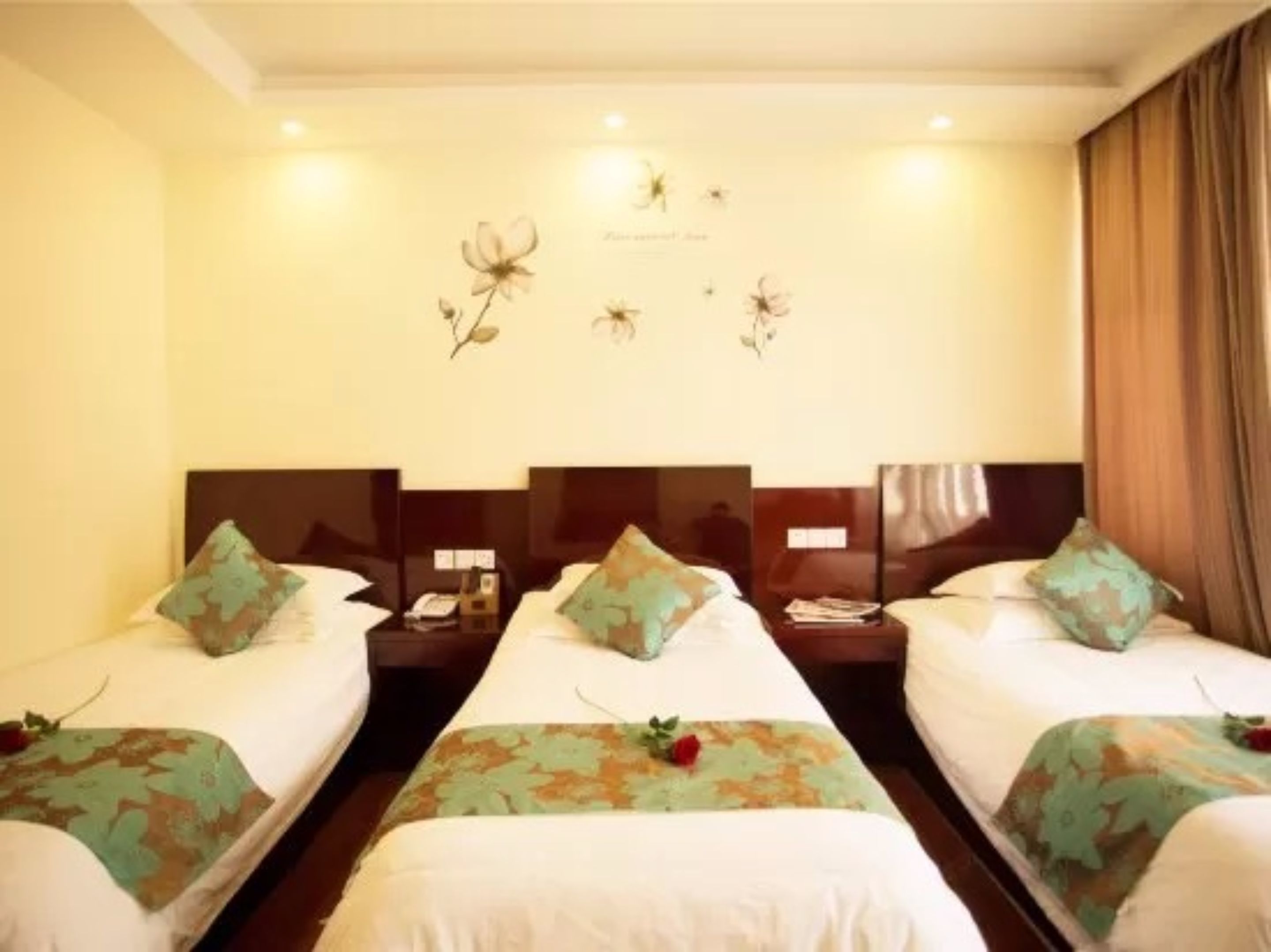 Ein Hotelzimmer mit drei Einzelbetten, jedes mit einer blumigen Tagesdecke und passenden Kissen. An jedem Bett steht ein Nachttisch mit einer Lampe und einem Telefon. Das Zimmer hat ein großes Fenster mit Vorhängen, und die Wände sind mit einem floralen Wandgemälde verziert.