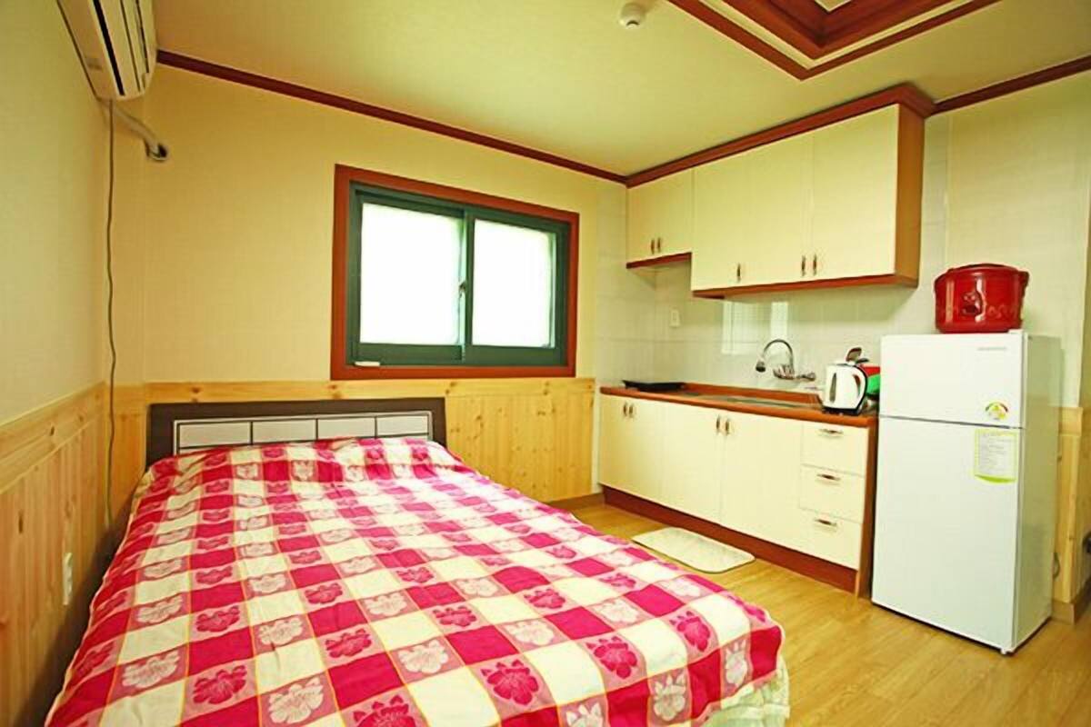 A bedroom with a bed, a window, a kitchen area with cabinets and a refrigerator.