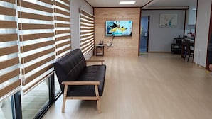 A modern living room with a black leather sofa, wooden blinds, a flat-screen TV on the wall, and a dining area in the background. - Blue Sky Pension (Pocheon)