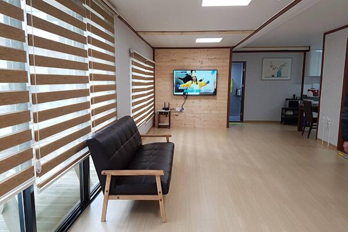 A modern living room with a black leather sofa, wooden blinds, a flat-screen TV on the wall, and a dining area in the background.