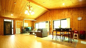 A spacious living area with a wooden floor, a fireplace, a dining table with chairs, and a large window providing natural light. - Blue Sky Pension (Pocheon)