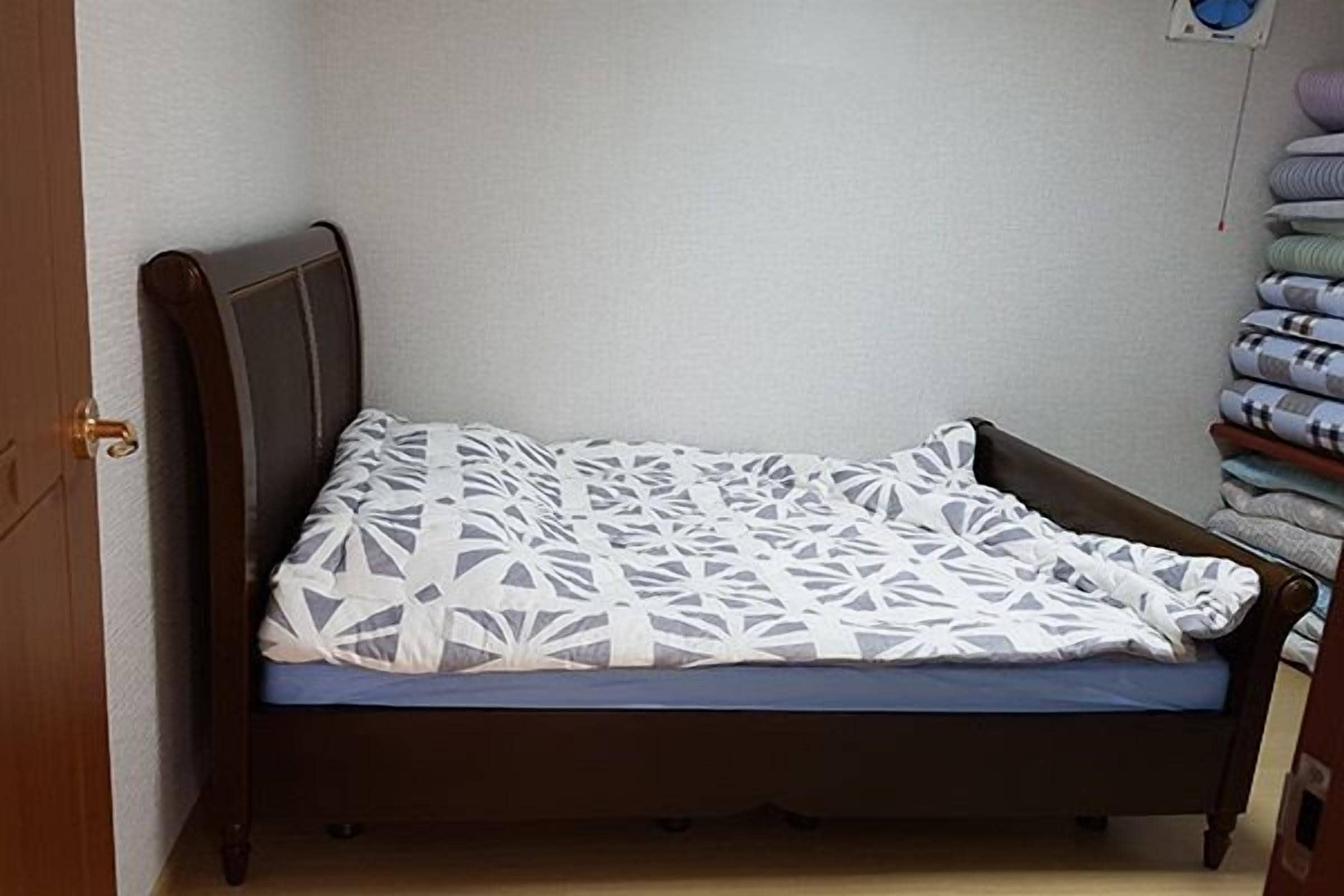 Quarto pequeno com uma cama de solteiro, cabeceira de madeira, edredom estampado e pilhas de cobertores dobrados em uma estante.