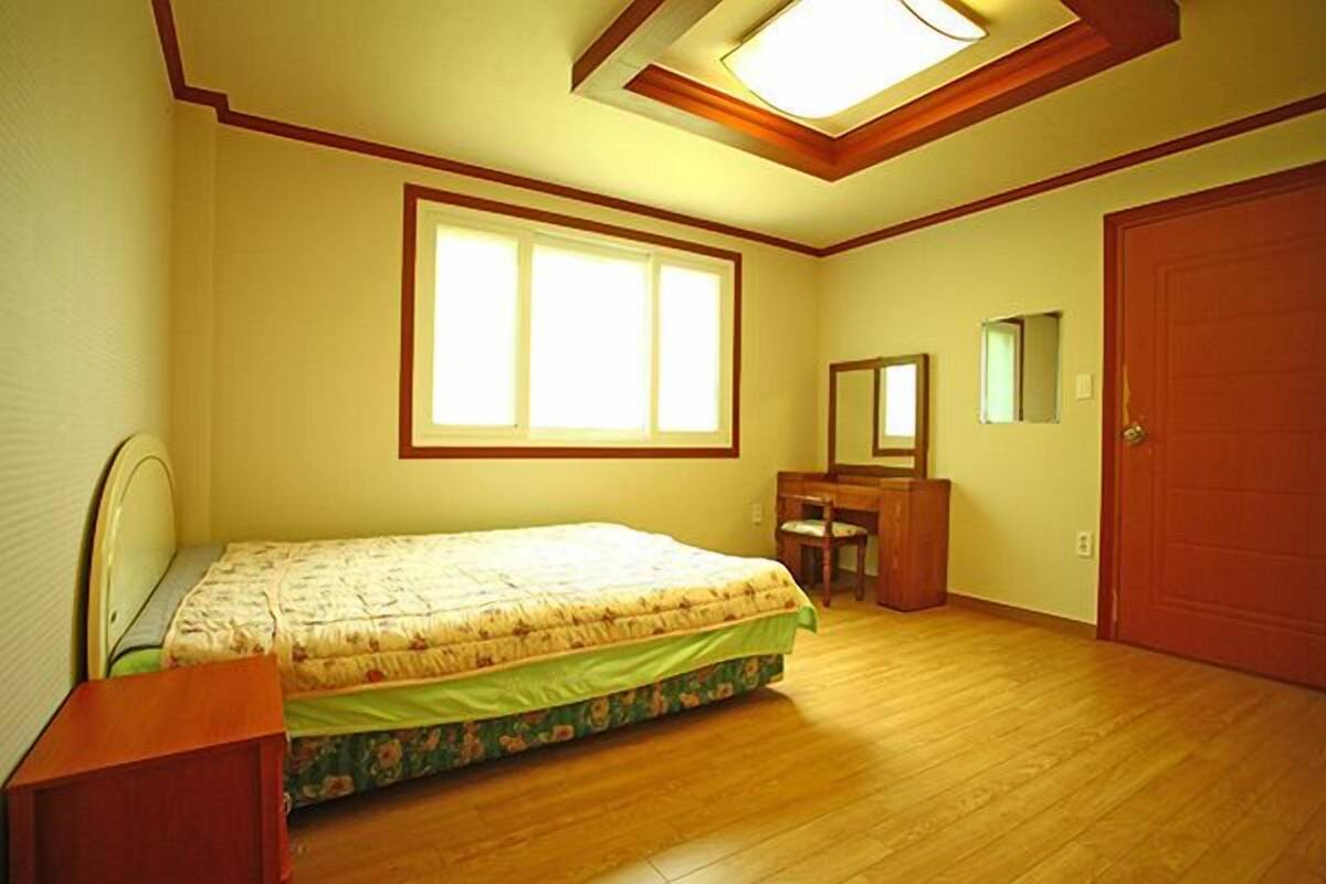 A bedroom with a bed, a desk, a chair, a mirror, and a door.