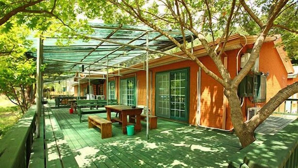 A covered outdoor area with wooden benches and picnic tables, surrounded by trees and a building with large windows. - Blue Sky Pension (Pocheon)