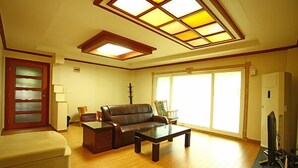A living room with a sofa, coffee table, and a TV. There is a window with blinds, a door, and a ceiling with skylights. - Blue Sky Pension (Pocheon)