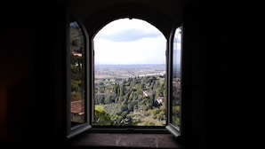 Une vue à travers une fenêtre dotée d’un cadre en arc de cercle, donnant sur un paysage arboré avec des bâtiments situés au loin.