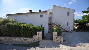 A two-story white building with a stone staircase leading to the entrance, surrounded by greenery and a stone wall. - Comfortable apartment with balcony (Pag)