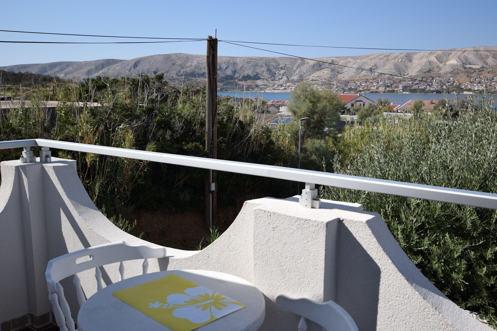 A balcony with a view of a lake and mountains, featuring a white railing and a table with a yellow placemat.