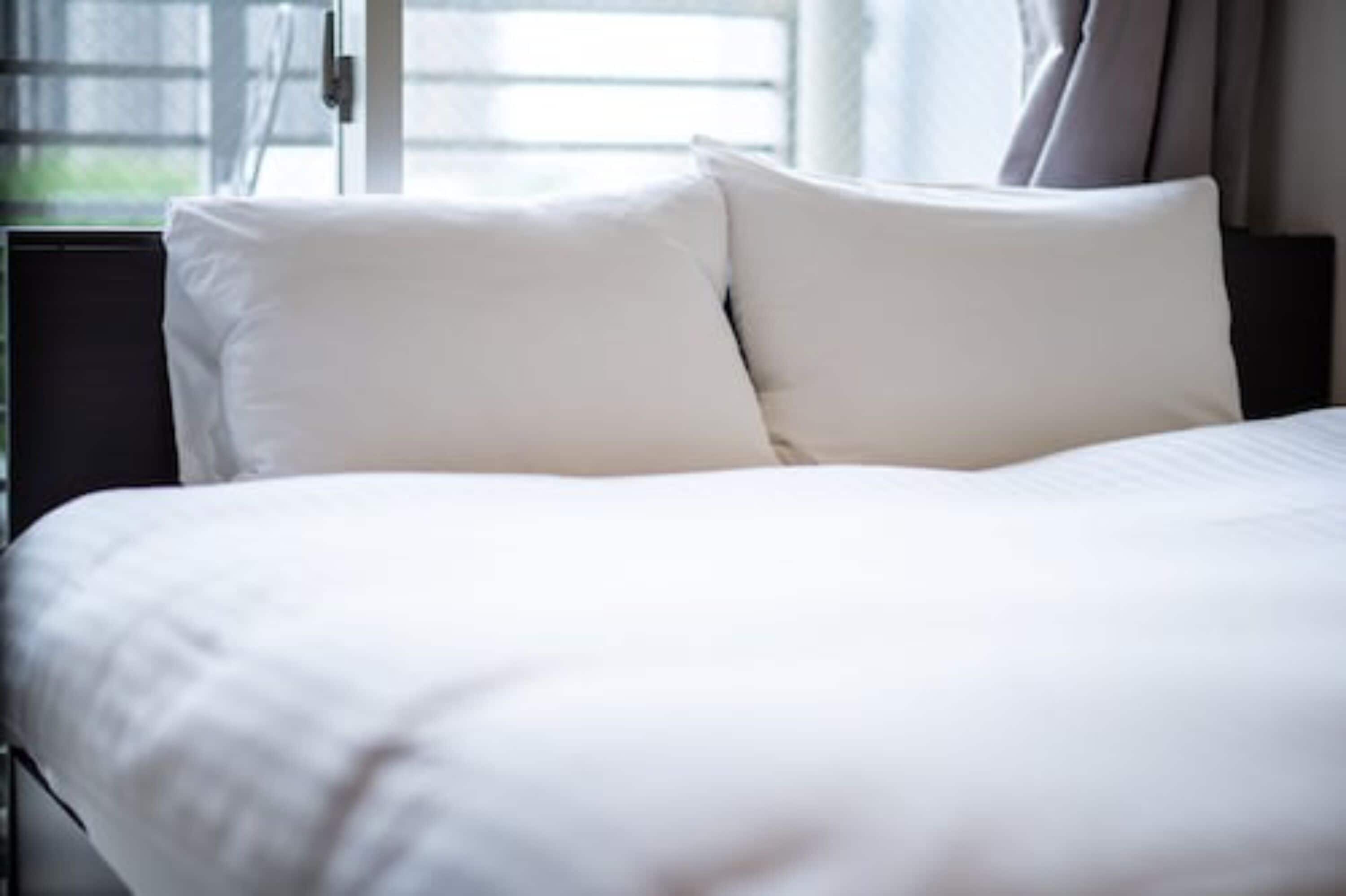 Een netjes opgemaakt bed met witte lakens en kussens, een donker hoofdbord en een raam met gordijnen op de achtergrond.