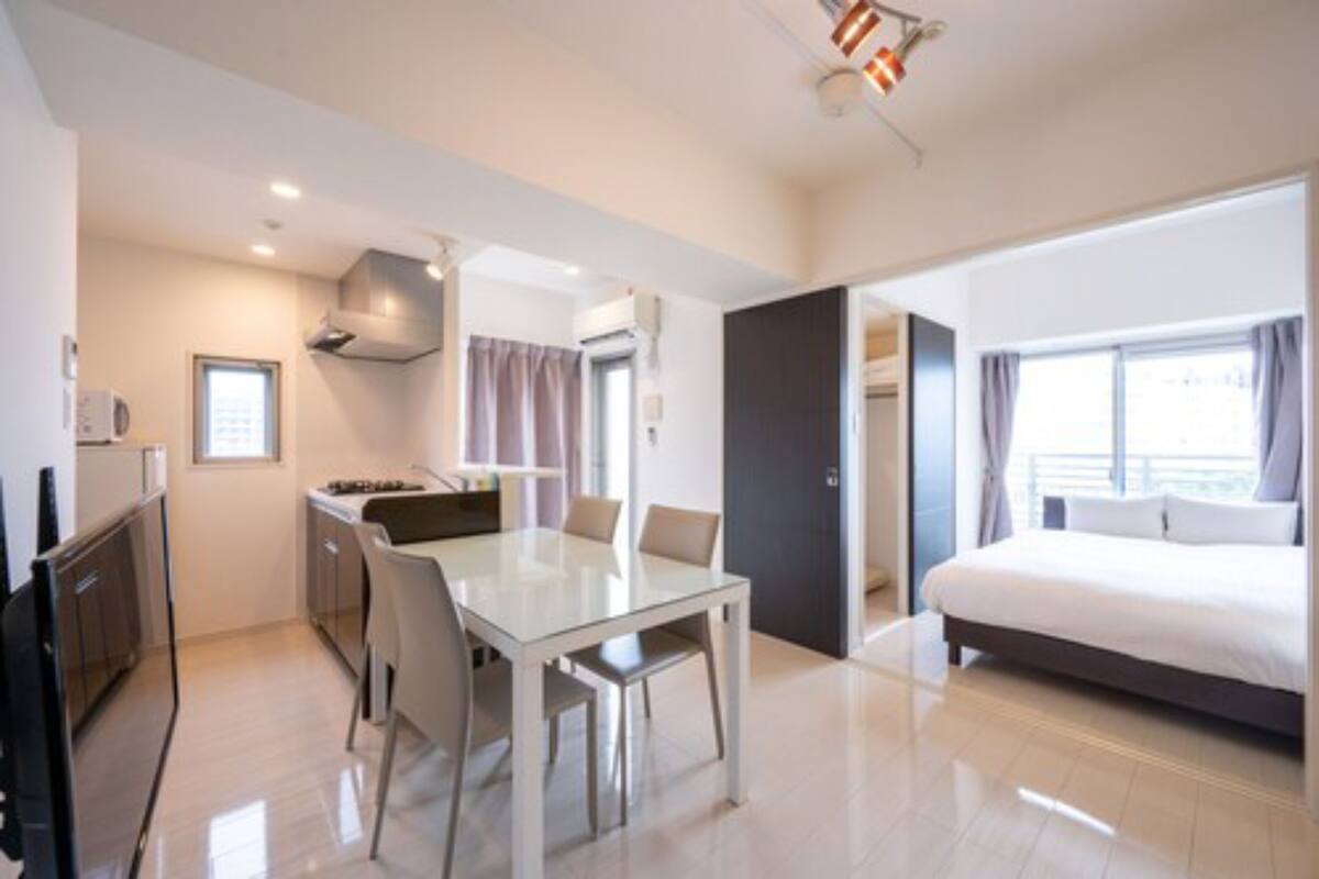 A modern hotel room with a bed, a dining area with a table and chairs, a kitchenette, and a large window with curtains.
