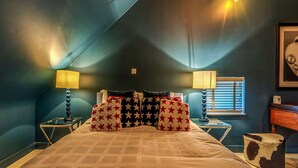 Ein Schlafzimmer mit schrägverdeckter Decke, einem Bett mit gemustertem Überwurf, zwei Nachttischen mit Lampen, einem Fenster mit Jalousien und einem gerahmten Bild an der Wand.