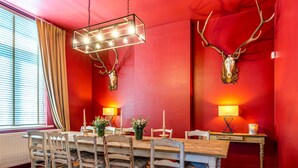 Ein Essbereich mit roter Wand, einem Holztisch und Stühlen. Zwei Wandleuchten und eine Deckenleuchte sind vorhanden.