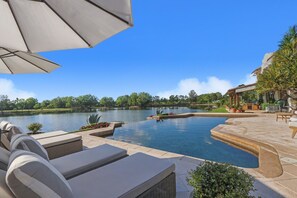 A swimming pool with lounge chairs and umbrellas, surrounded by a landscaped garden and a view of a lake. -  One of A Kind Waterfront Retreat with Scenic Views & Designer Interiors (Jupiter)