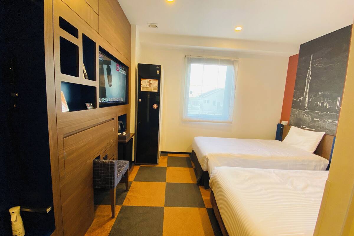 Ein Hotelzimmer mit zwei Betten, einem Fernseher, einem kleinen Tisch, einem Stuhl, einem Fenster und einer Tür.